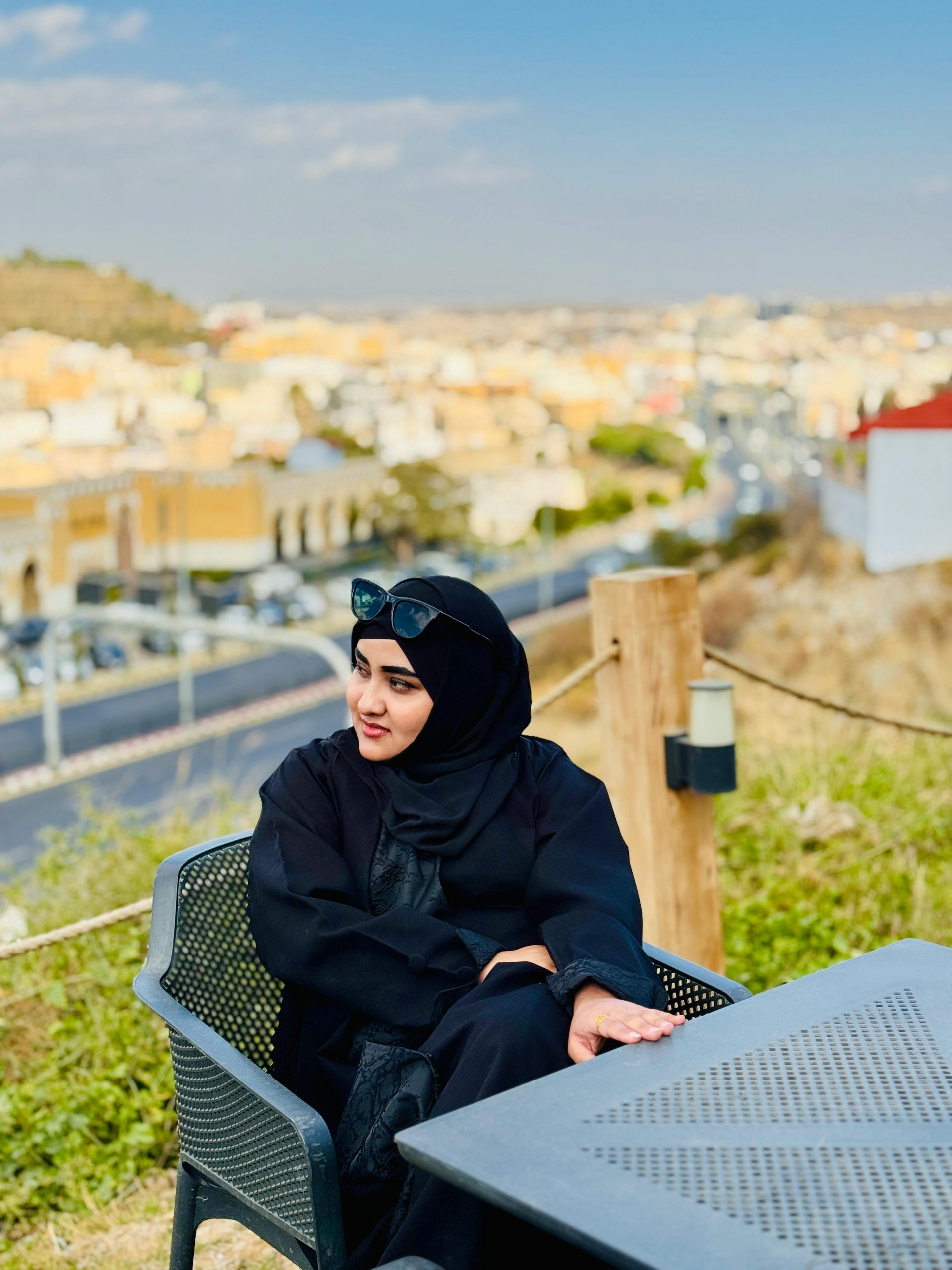 Woman in black abaya seated outdoors with scenic urban view, sunny day.