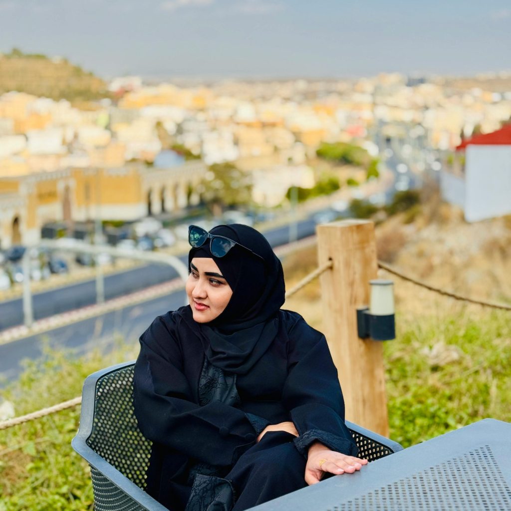 Woman in black abaya seated outdoors with scenic urban view, sunny day.